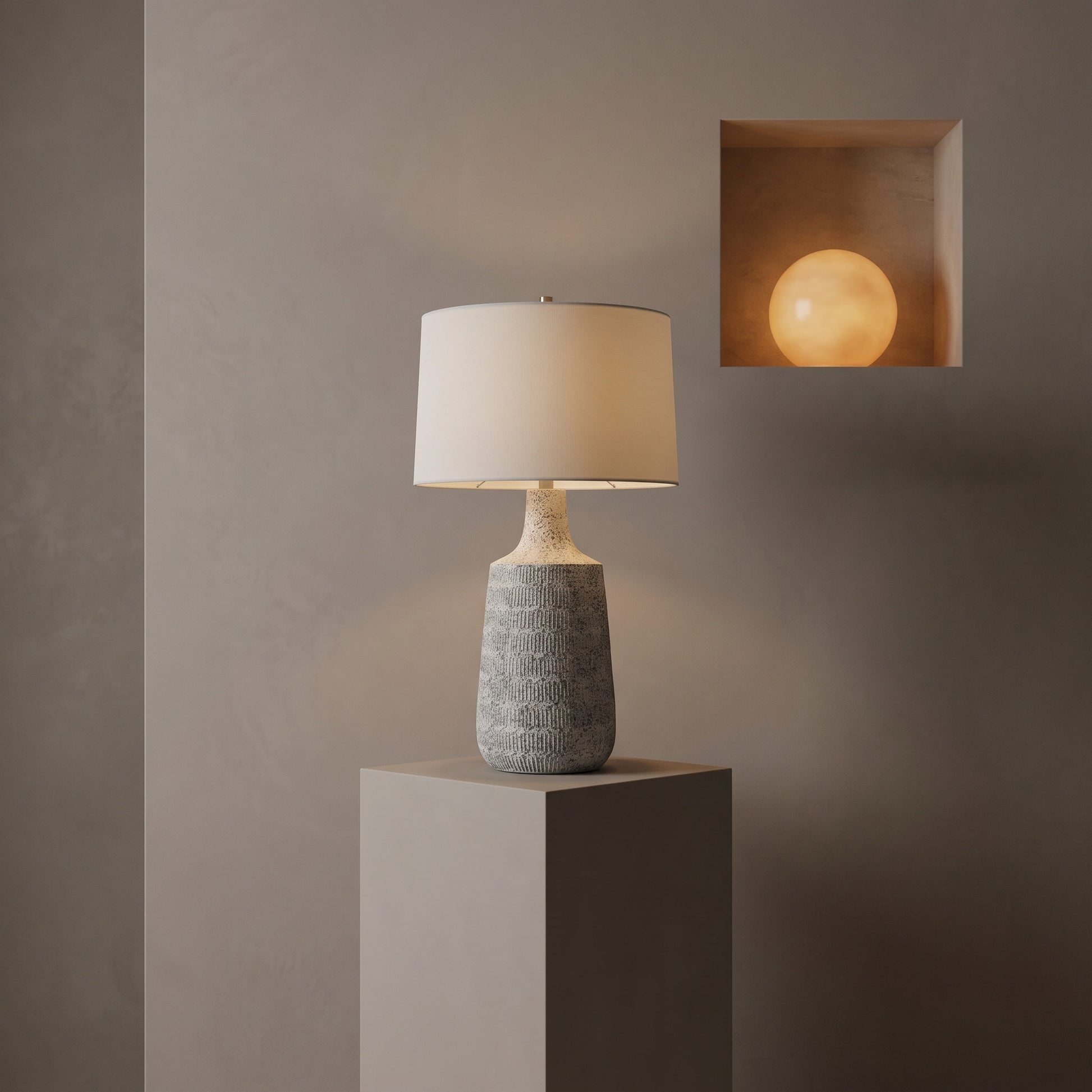 A table lamp with a textured gray ceramic base and a warm off-white drum shade, displayed on a gray pedestal against a textured gray wall. A recessed wall light in the upper right glows with a warm yellow light.