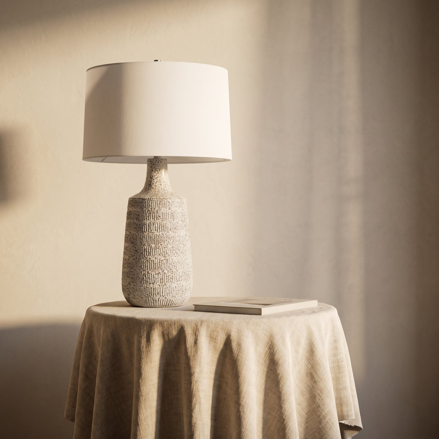 A table lamp with a textured beige ceramic base and a white drum shade, styled on a draped beige tablecloth next to a book against a warm beige wall.