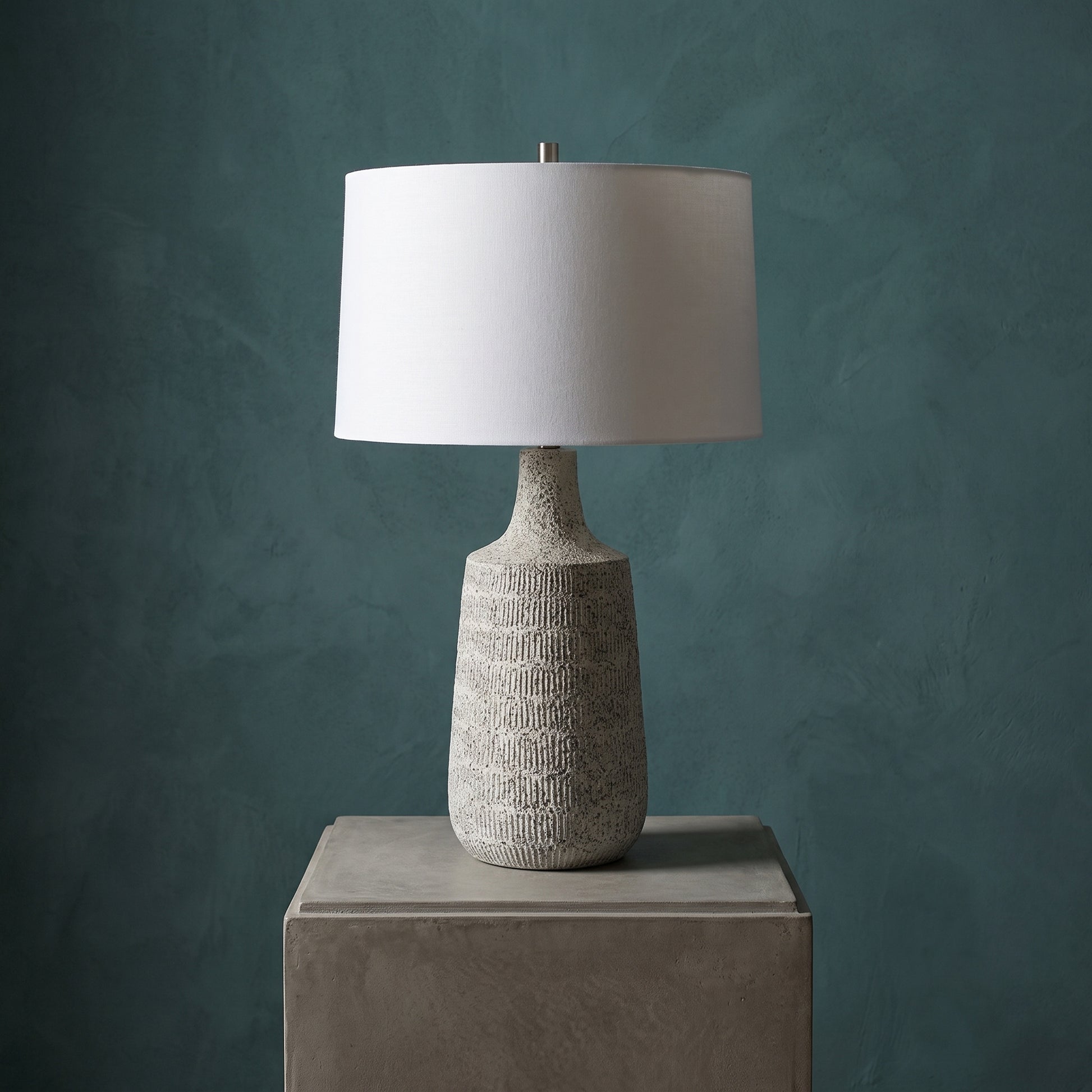A table lamp with a textured gray ceramic base and a white drum shade, displayed on a matching gray pedestal against a dark teal textured wall.