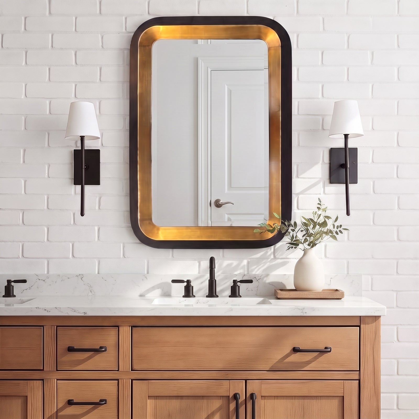A rectangular wall mirror with a black frame and warm gold inner trim, mounted above a double-sink bathroom vanity with a white brick wall and two black sconces.
