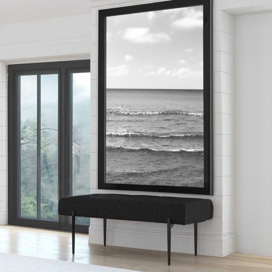 White bedroom overlooking forest with hardwood floors and photograph of ocean with a black bedroom bench.