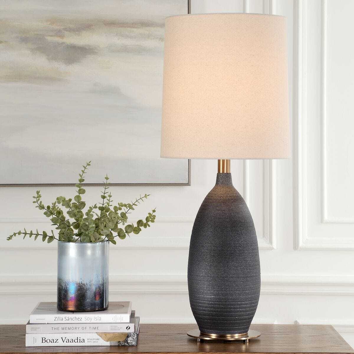 Decorative table lamp with a textured base and beige shade on a wooden surface, with books and a plant in the background.