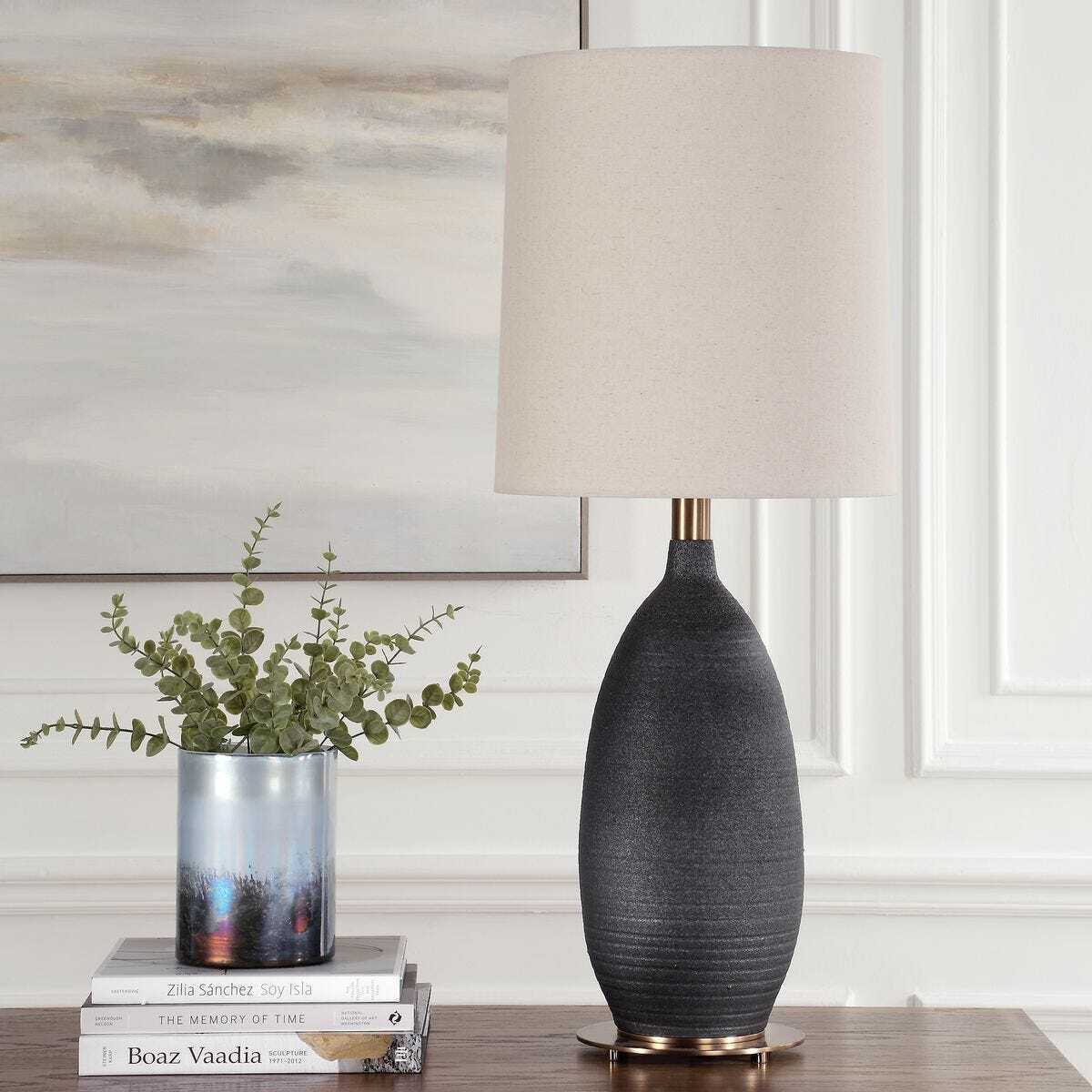 Gray textured table lamp with beige shade on a wooden surface, with books and a plant in the background.
