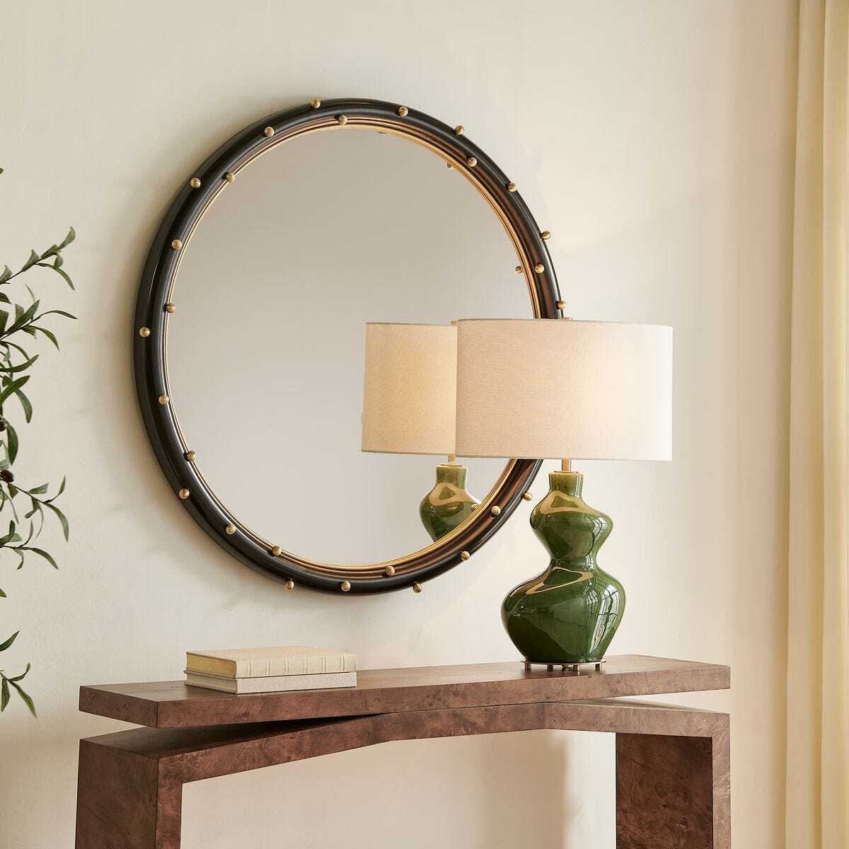 Round mirror with decorative frame on a wall above a wooden console table with a green lamp.