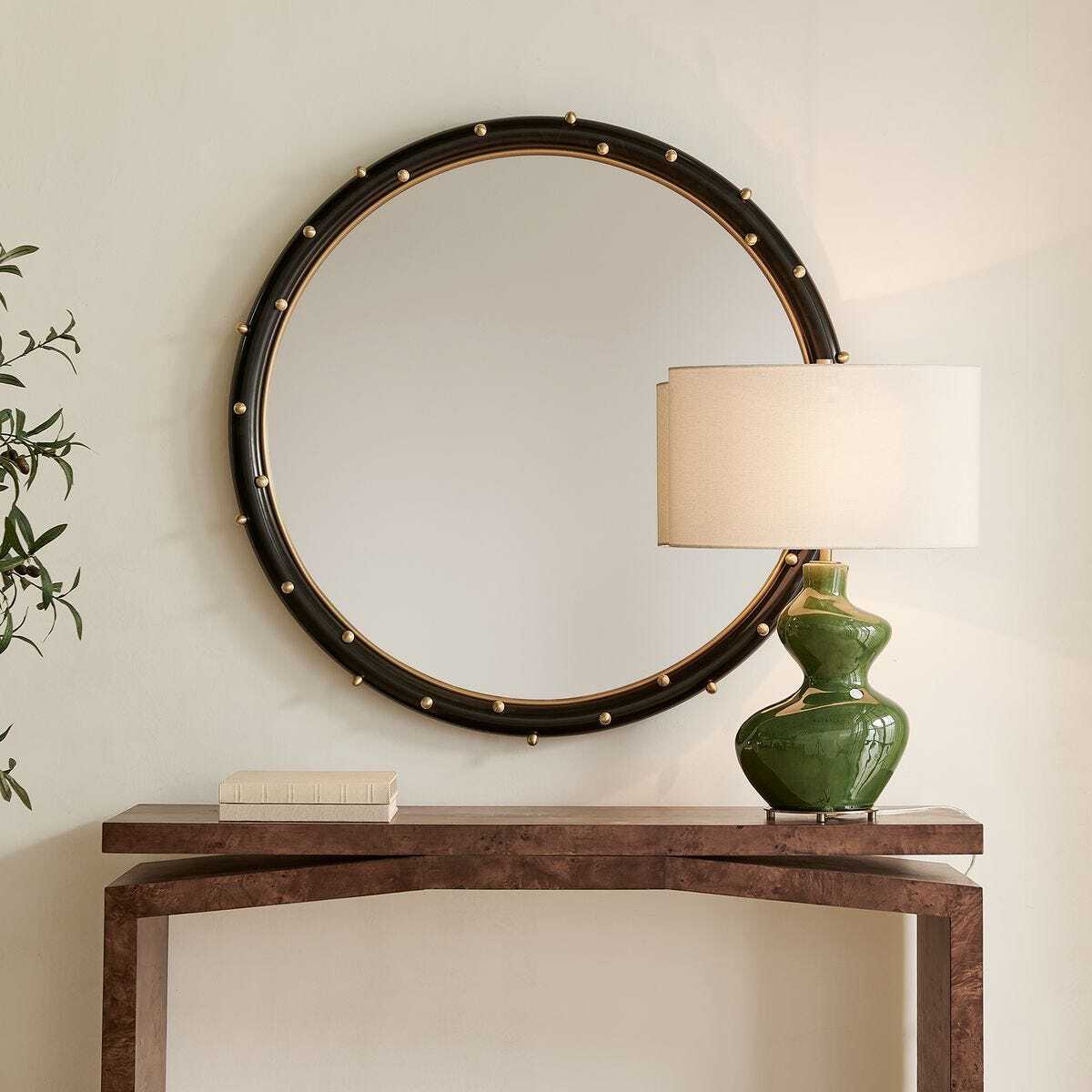 Round mirror with decorative frame above a wooden console table with a green lamp.
