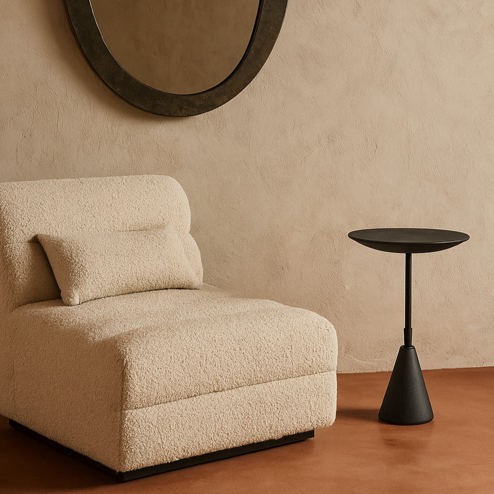 Beige armchair and black side table against a beige wall with a round mirror.