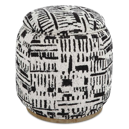 A round upholstered pouf ottoman with a black and white abstract ikat-style pattern, set on a textured beige rug in a bright room.