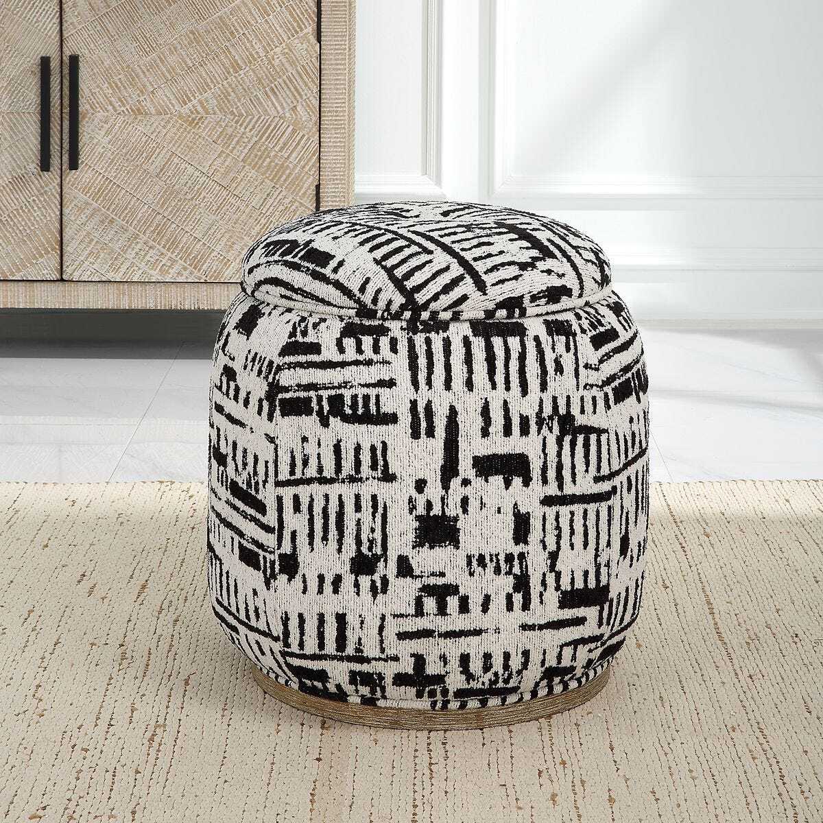 A round upholstered pouf ottoman with a black and white abstract ikat-style pattern, set on a textured beige rug in a bright room.