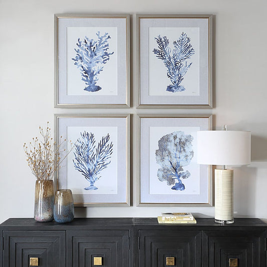 Four painting of blue coral wall art above a black wood console with gold knobs and a white coastal lamp.