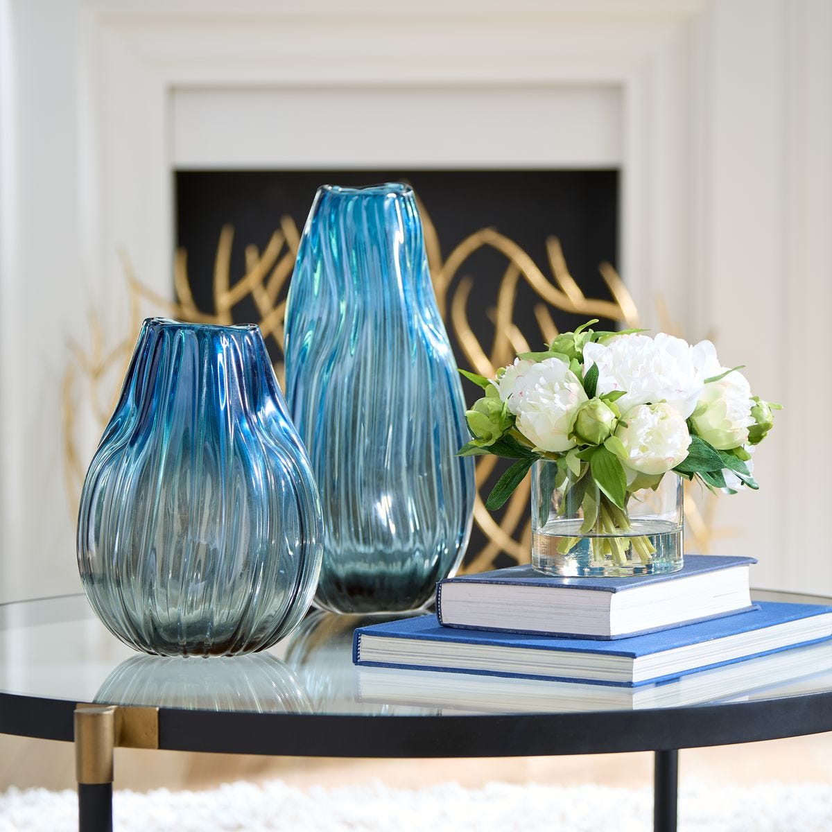 Two blue glass vases on a table with a small vase of white flowers and books in the background.