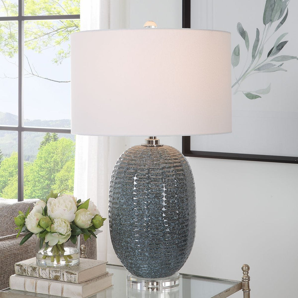 Decorative table lamp with textured gray base and white shade in a room with a window and plant.