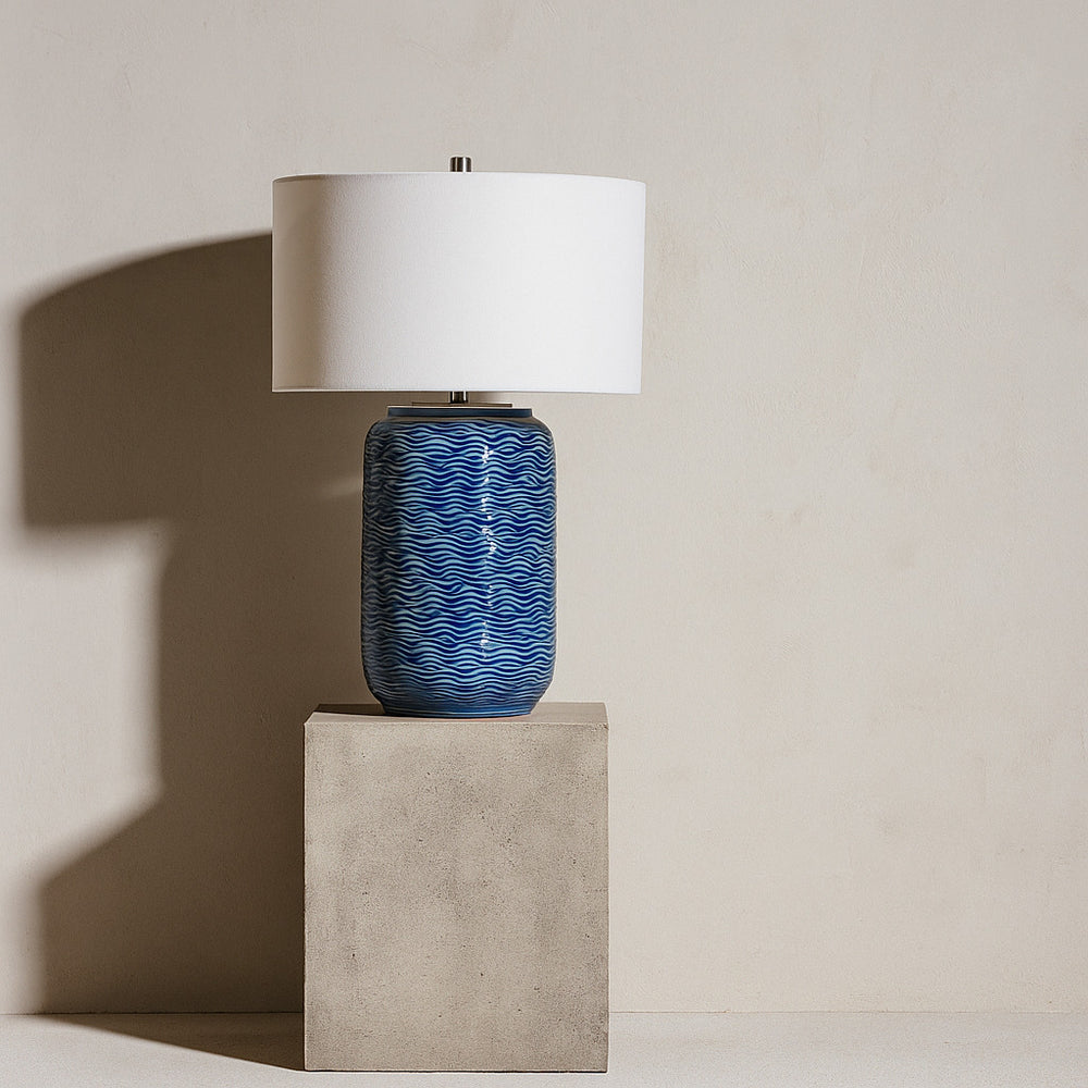 Table lamp with blue textured base and white shade on a beige pedestal against a beige wall.