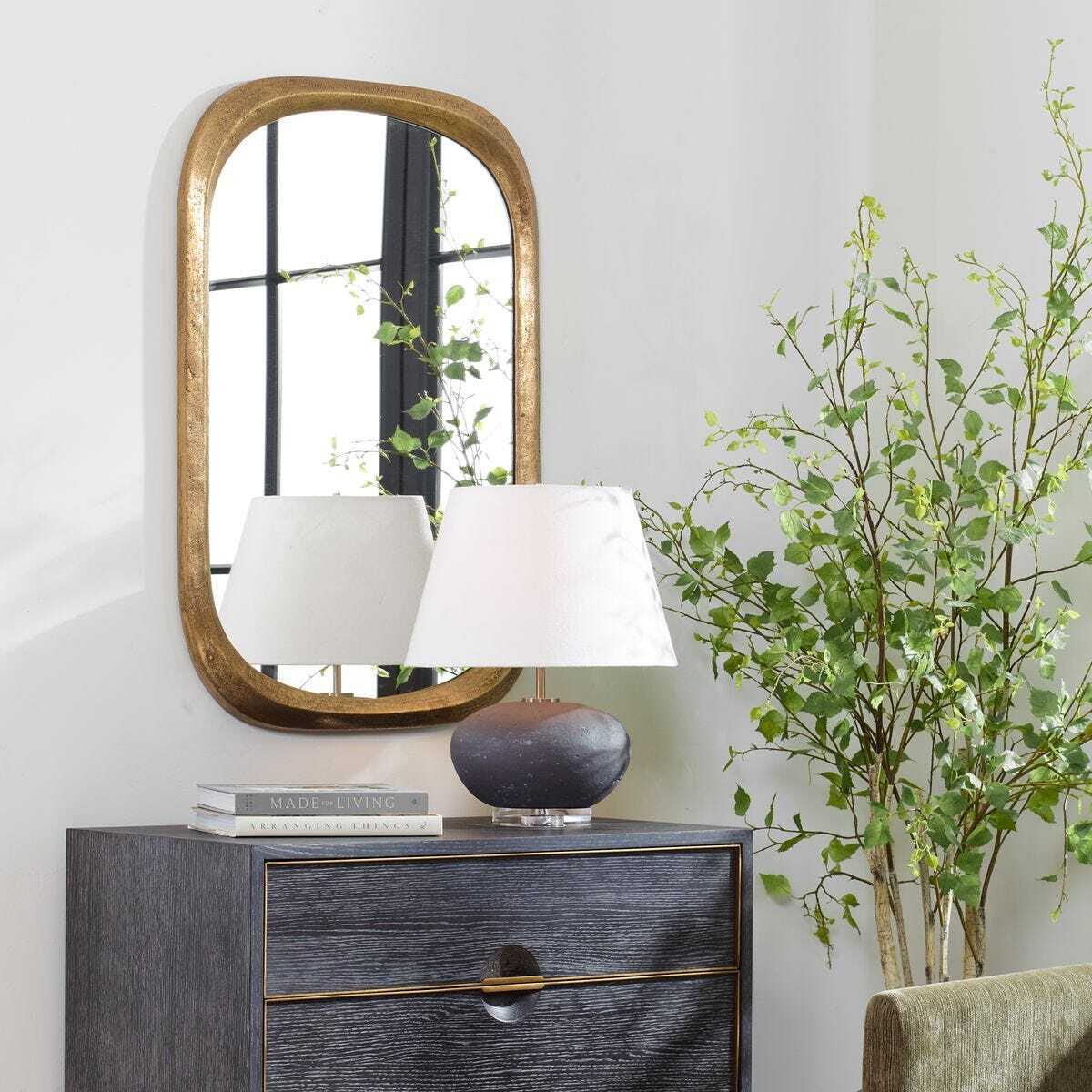 Decorative mirror on a side table with a lamp and books, next to a plant against a white wall.