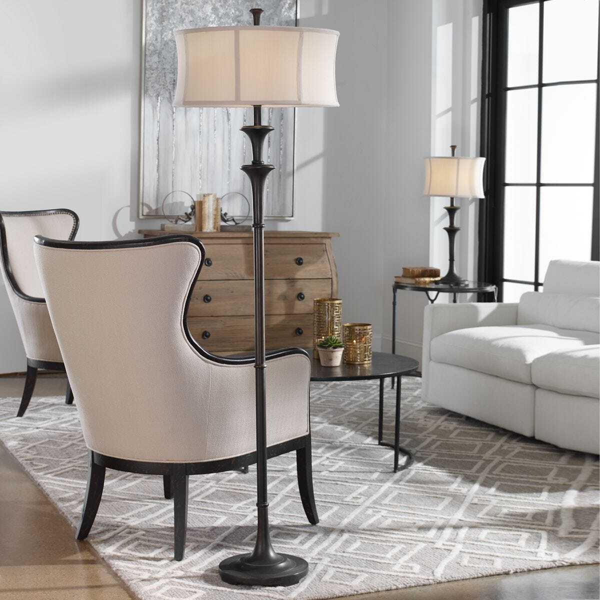 A modern living room scene featuring a tall floor lamp with a black metal frame and a cream fabric shade, positioned next to a beige upholstered wingback armchair. The room is styled with a white sofa, a wooden chest, and a patterned rug.