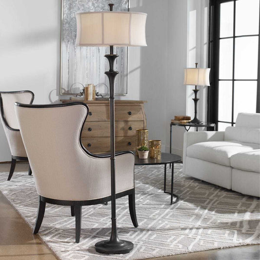 A floor lamp with a dark bronze metal stand and a cream fabric drum shade, shown in a minimalist white room corner with its shadow cast to the right.