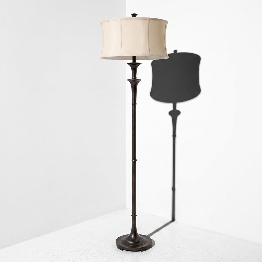 A floor lamp with a dark bronze metal stand and a cream fabric drum shade, shown in a minimalist white room corner with its shadow cast to the right.