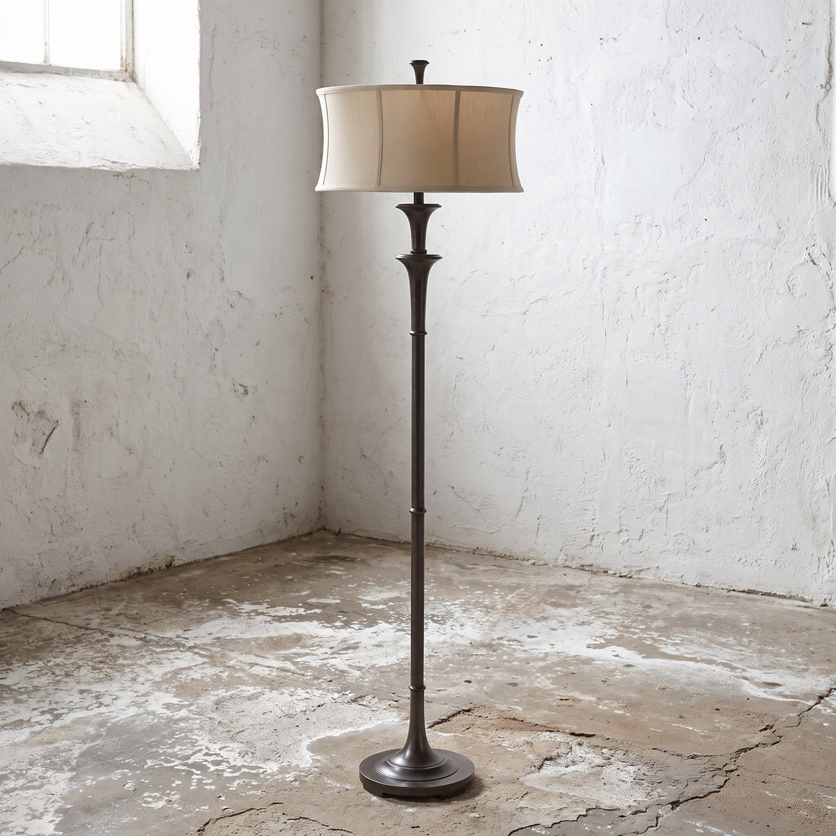 A tall floor lamp with a dark bronze metal stand and a beige fabric drum shade, positioned in the corner of a rustic room with white plaster walls and a concrete floor.