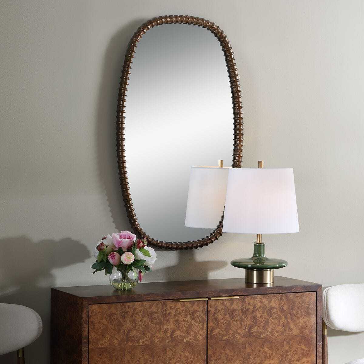Wooden dresser with oval mirror, lamp, and flowers against a beige wall.