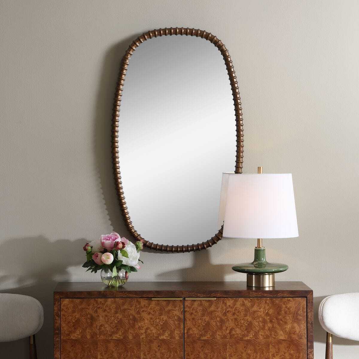 An oval wall mirror with a dark beaded frame hangs above a wooden console table in a styled interior. A lit table lamp with a white shade and a vase of pink flowers sit on the table against a light neutral wall.
