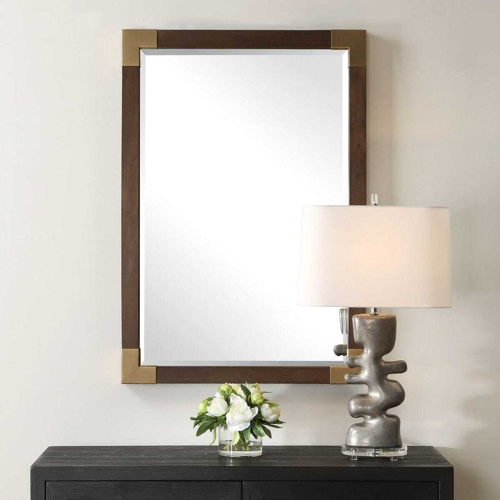 Decorative mirror with wooden frame above a table with a lamp and floral arrangement.