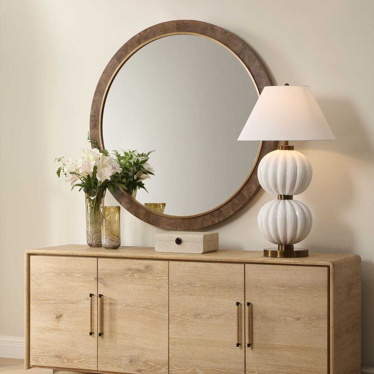 Wooden sideboard with round mirror, lamp, and flowers against a beige wall.