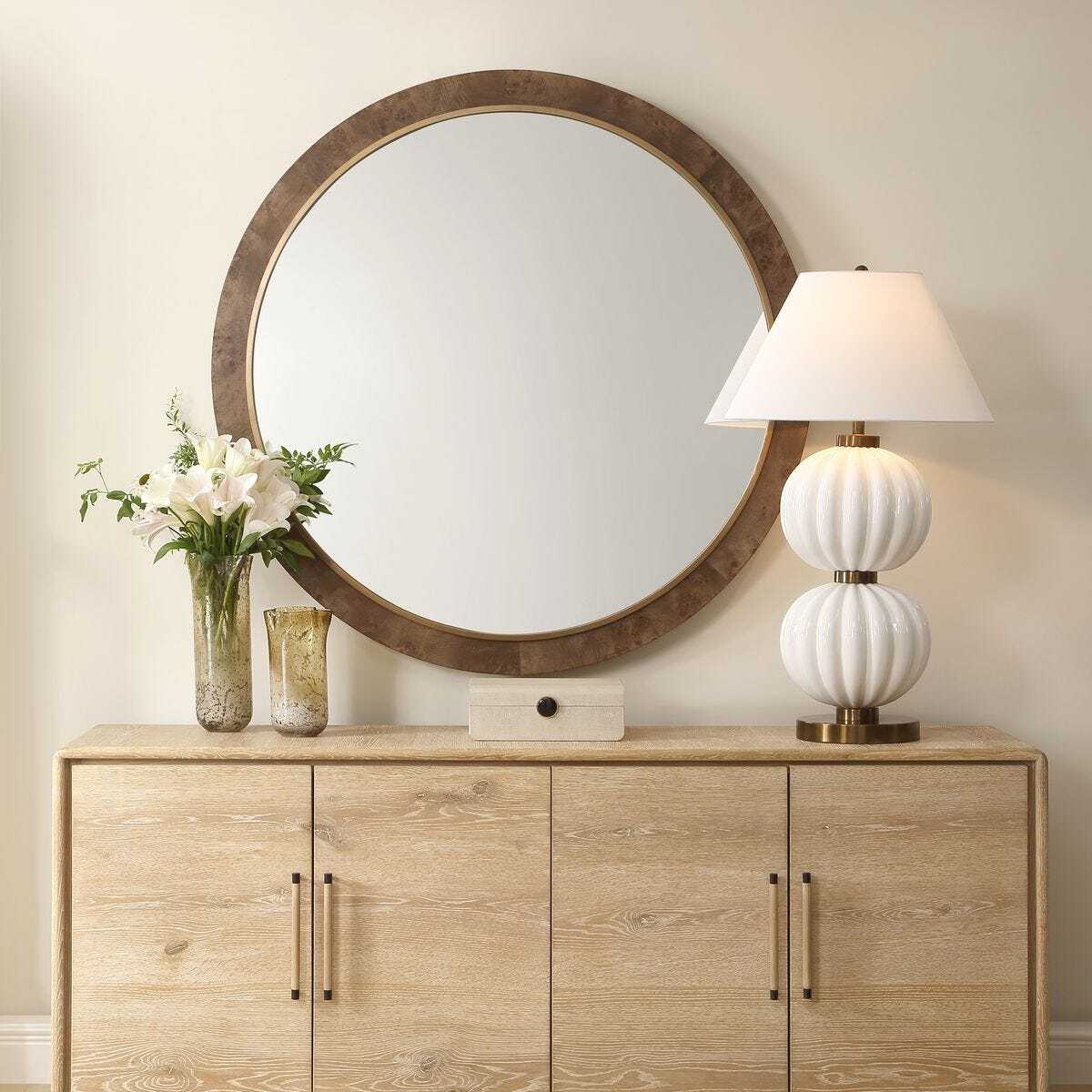 Wooden sideboard with round mirror, lamp, and flowers against a beige wall.