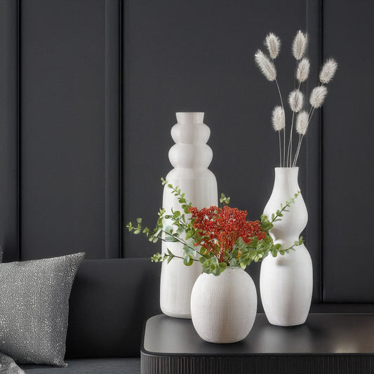 three ceramic white vases with flowers on black end table with black walls.