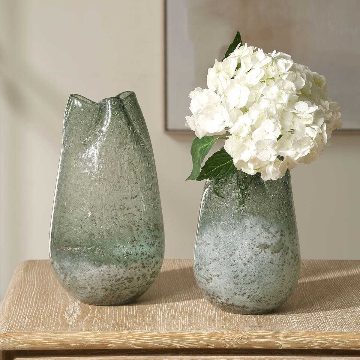 Two green glass vases on a wooden surface with one containing white flowers.