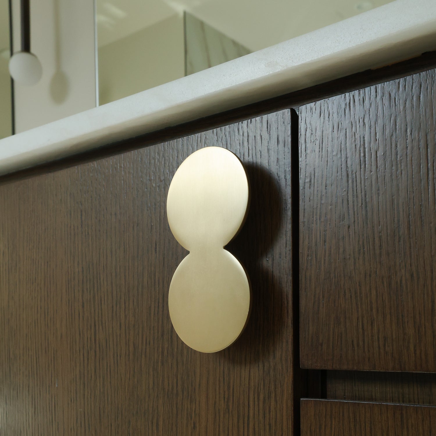 A close-up of a modern double-oval brushed brass cabinet pull handle mounted on a dark brown wood cabinet door.