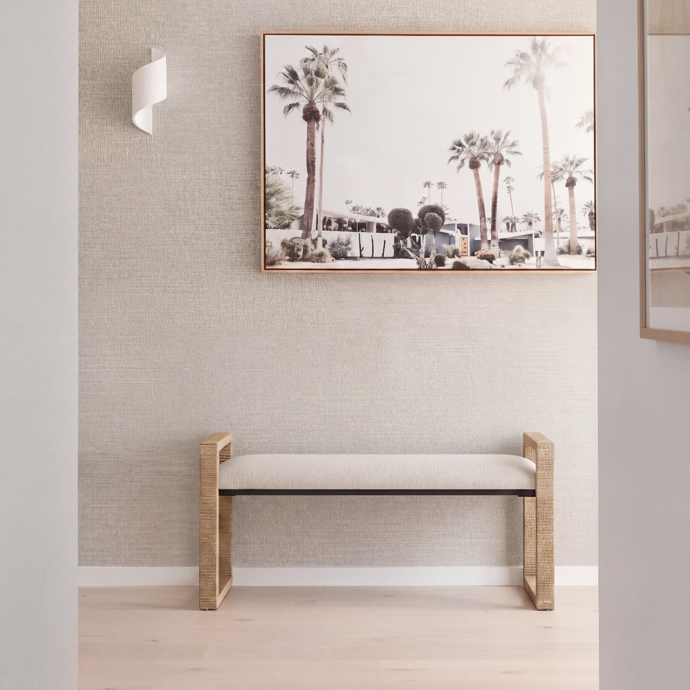 A modern wooden bench with a light beige upholstered seat is placed against a textured light gray wall in a bright interior. Above the bench hangs a framed art print depicting palm trees.