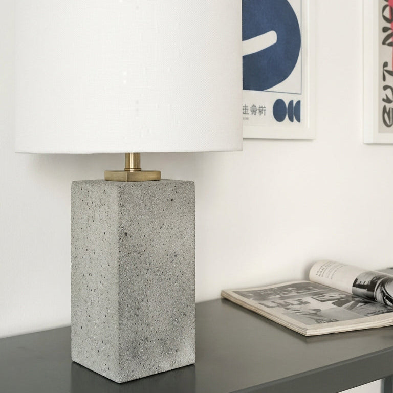 Table lamp with concrete base on a table next to a newspaper, with abstract art in the background.