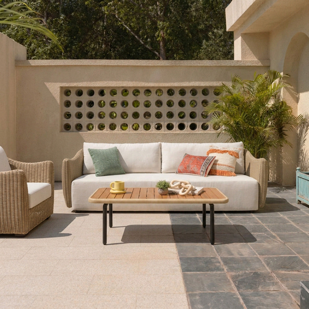 Outdoor patio furniture set featuring a beige sofa with white cushions and decorative pillows, a matching armchair, and a wooden slat-top coffee table on a tiled patio in front of a stucco wall with circular cutouts.