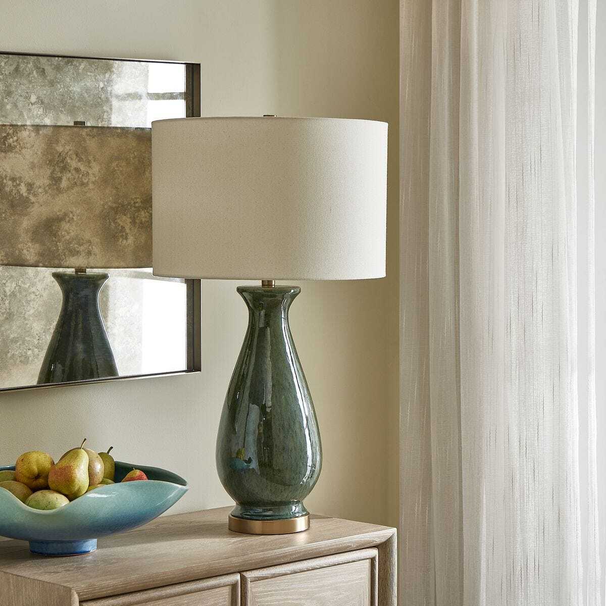 Decorative lamp with a white shade on a wooden surface, with a mirror and fruit bowl in the background.