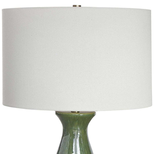 Decorative lamp with a white shade on a wooden surface, with a mirror and fruit bowl in the background.