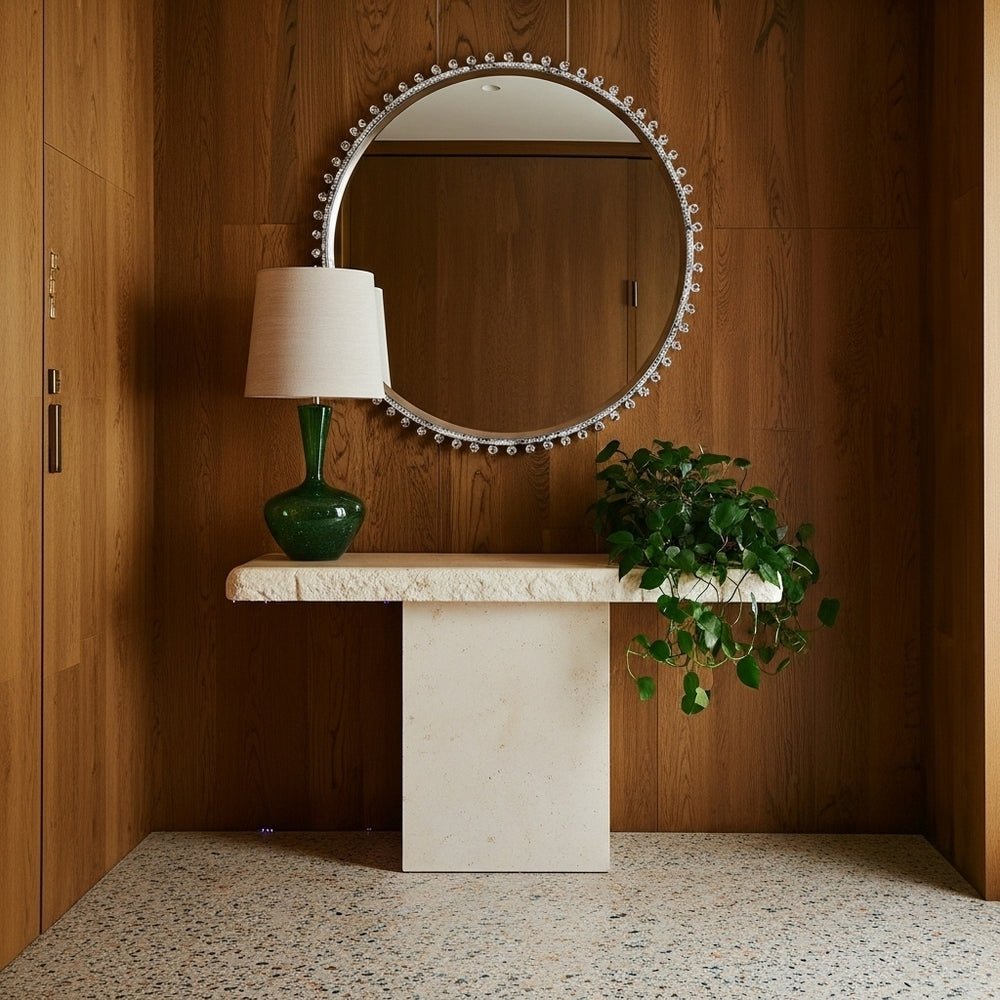 Round mirror with decorative frame above a marble console table with a lamp and plant against a wooden wall.