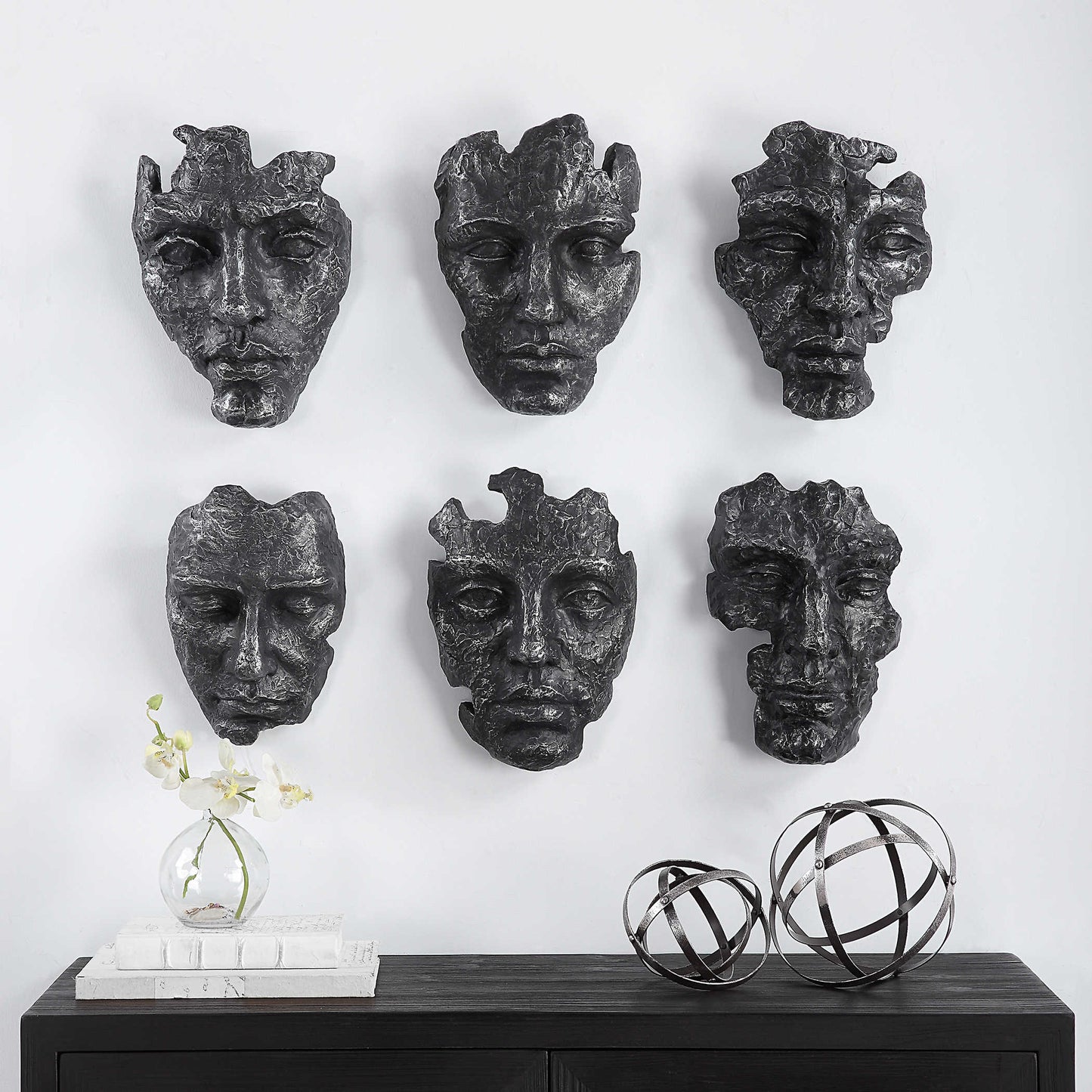 Set of six dark gray wall-mounted masks on a white wall with decorative items on a black surface below.