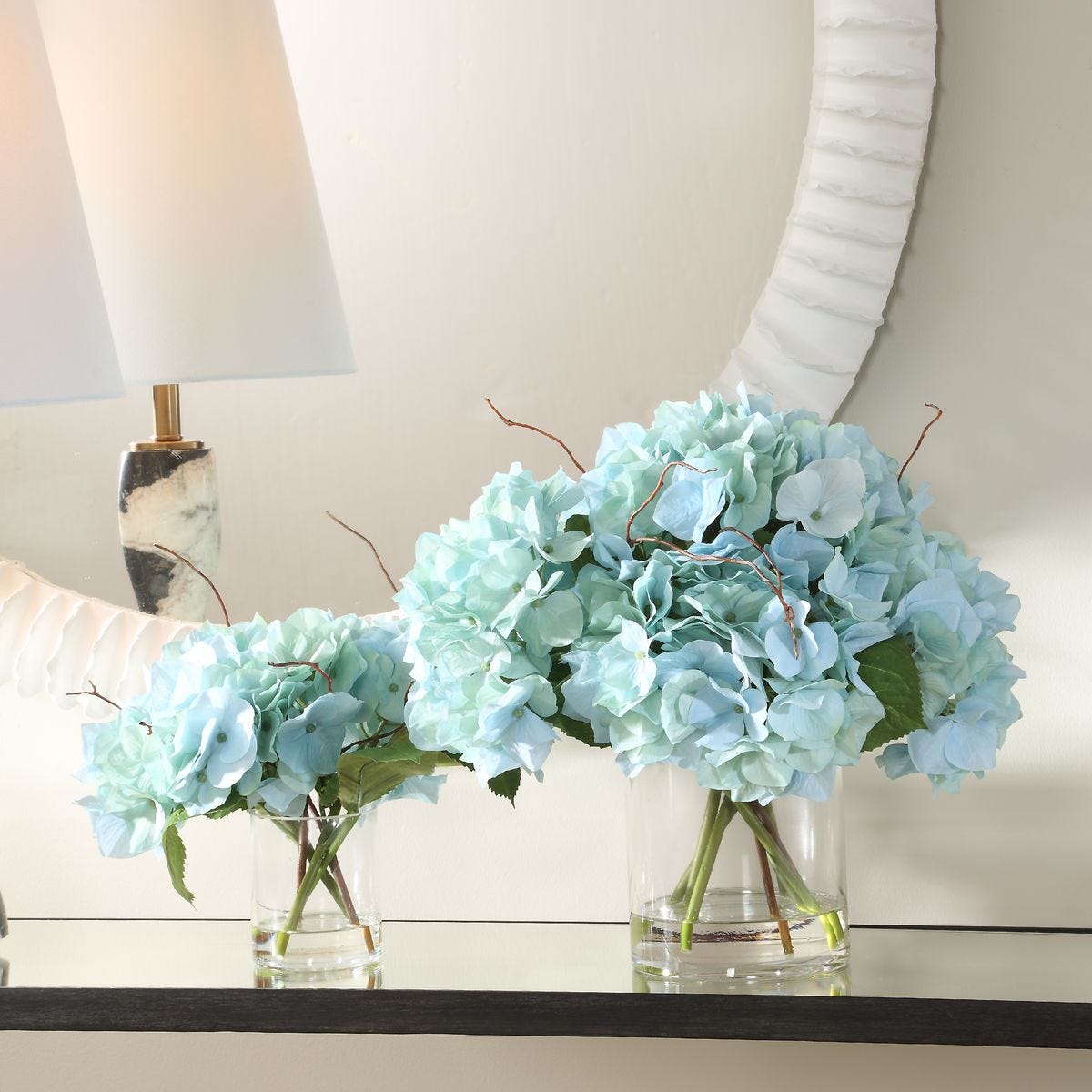 Light blue hydrangeas in clear vases on a table with a lamp and mirror in the background.