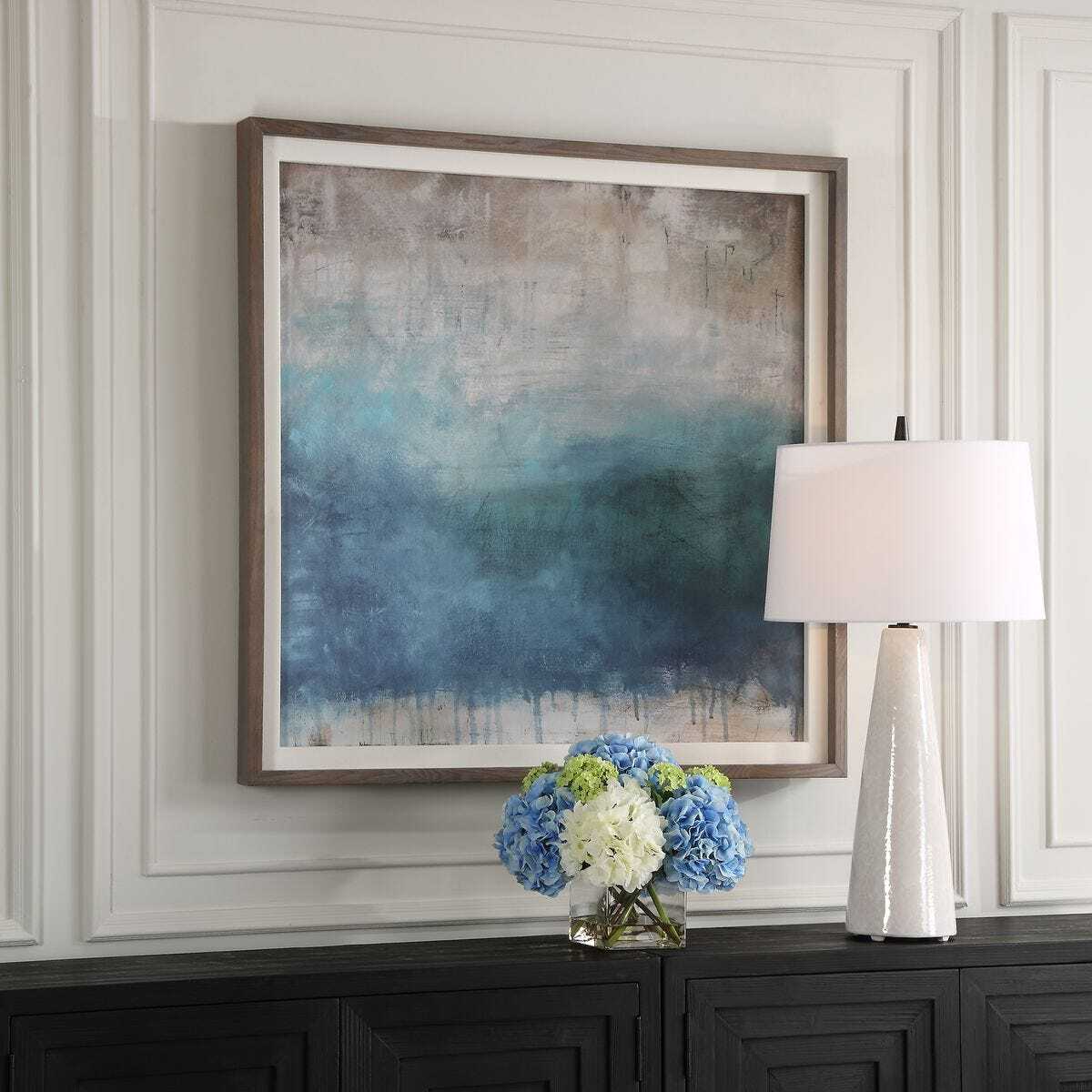 Abstract painting on a wall with a vase of flowers and a lamp on a dark console table.