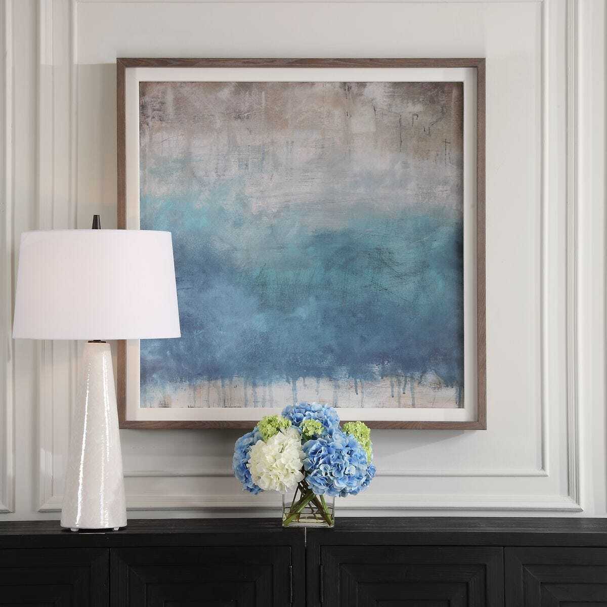 Framed abstract art piece on a wall above a console table with a lamp and floral arrangement.