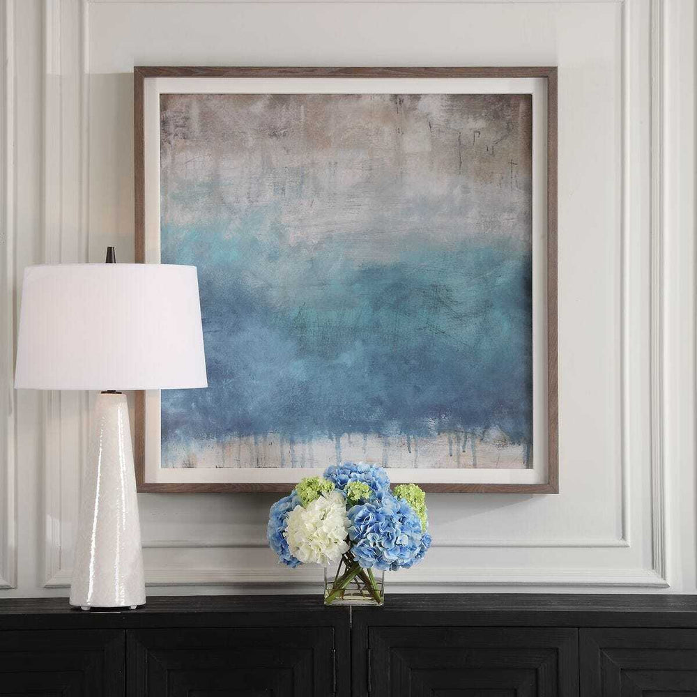 Framed abstract art piece on a wall above a console table with a lamp and floral arrangement.