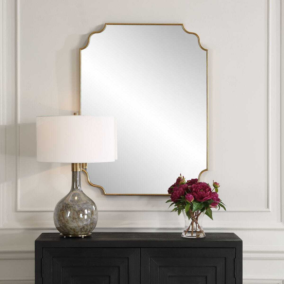 Decorative lamp and vase with flowers on a dark console table against a wall with a large mirror.