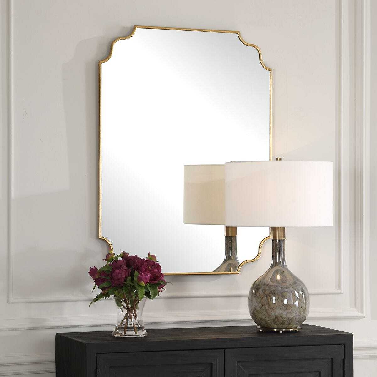 Decorative mirror with gold frame above a dark wooden cabinet with a lamp and flowers.