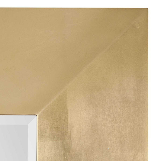 A large rectangular wall mirror with a wide, textured metallic gold frame, leaning against a neutral beige plaster wall on a light stone floor.