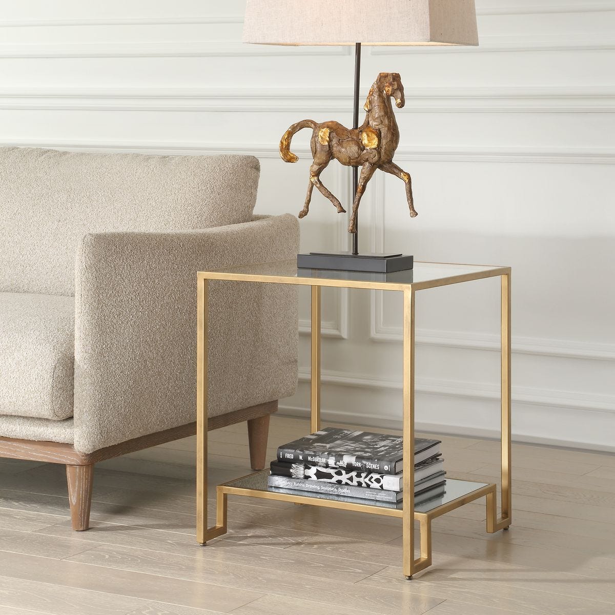 Gold side table with a lamp and books next to a beige armchair in a room.