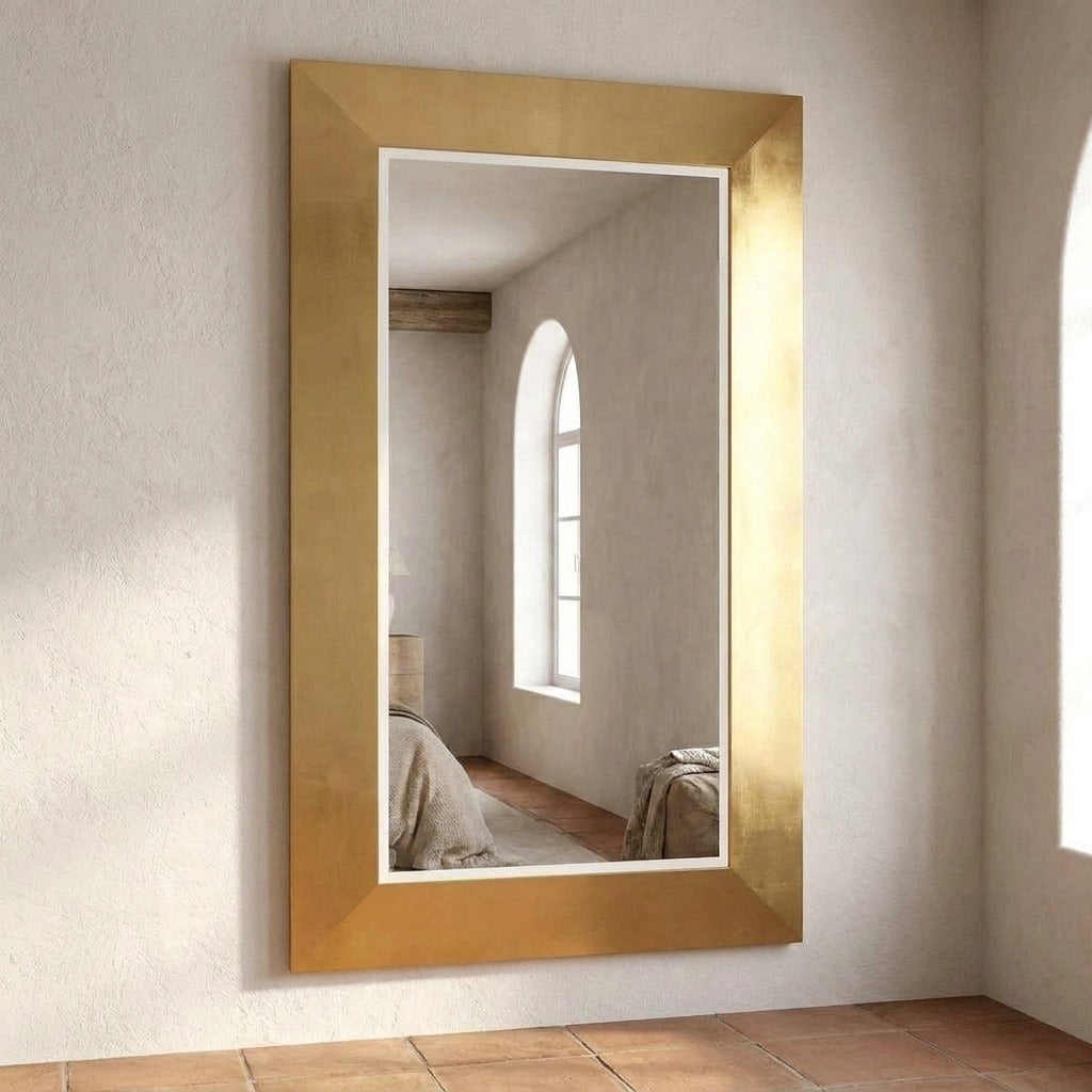 A tall rectangular wall mirror with a wide, textured gold frame, mounted on a light beige plaster wall above terracotta tile flooring.
