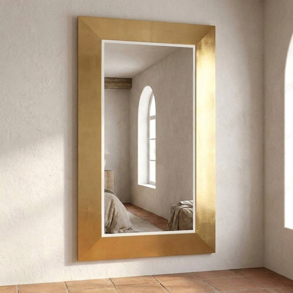 A tall rectangular wall mirror with a wide, textured gold frame, mounted on a light beige plaster wall above terracotta tile flooring.