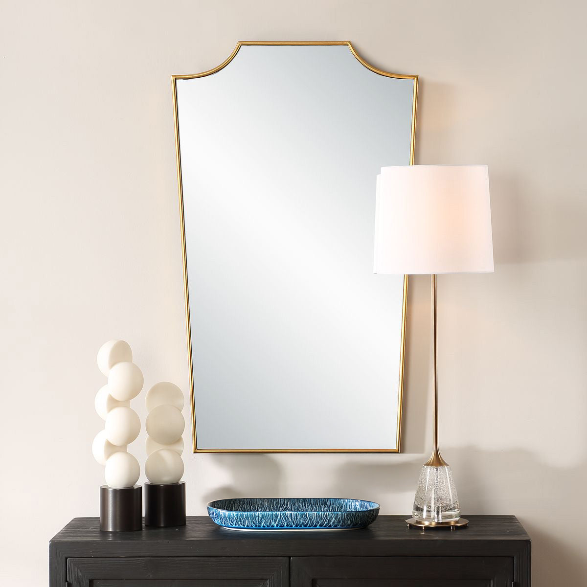 Fancy gold mirror hanging on wall above black dresser with white statue and blue tray and table lamp. 