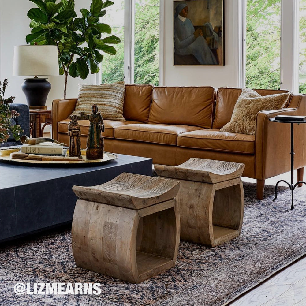 A styled living room scene featuring two rustic curved wooden stools in the foreground, a tan leather sofa, a dark coffee table, and a large potted fiddle-leaf fig in a bright room with tall windows.