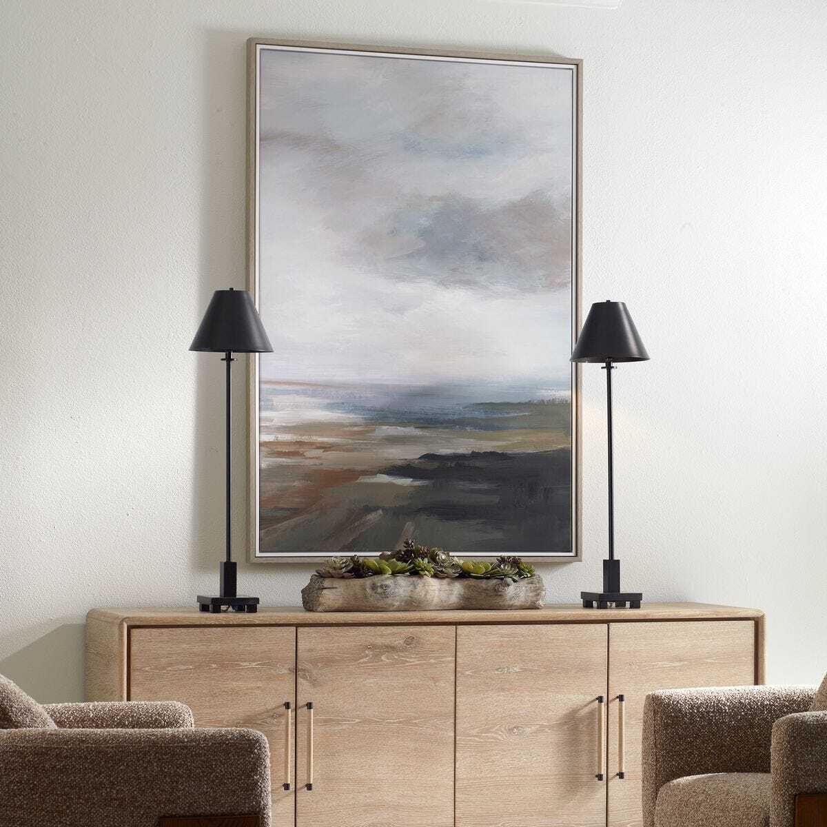 A large framed abstract landscape painting hangs on a white wall above a light wood sideboard, flanked by two black table lamps and a small planter. Two upholstered chairs are partially visible at the bottom corners.