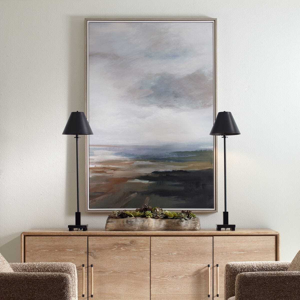 A large framed landscape painting with soft gray, blue, and brown tones hangs on a light wall above a wooden console table, flanked by two black table lamps and framed by the tops of two upholstered chairs.